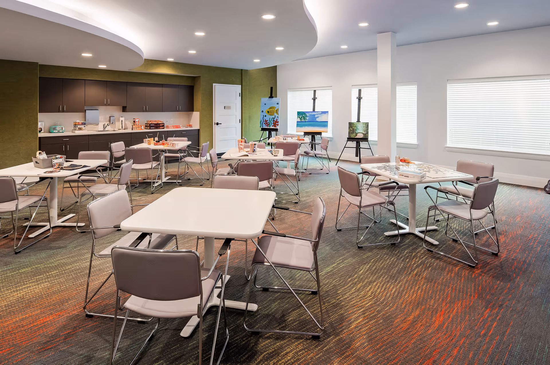 A bright and spacious activity room with multiple square tables and chairs arranged for group activities. The room features a colorful carpet, a kitchenette area with cabinets and a countertop along one wall, and three easels displaying paintings near the windows with blinds.
