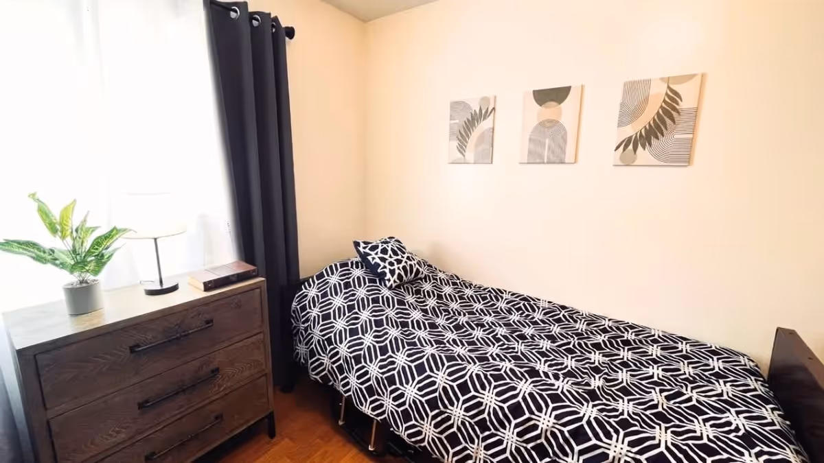 Small bedroom with a single bed covered in a black-and-white geometric comforter, a wooden dresser with a lamp and plant, and three pieces of wall art.