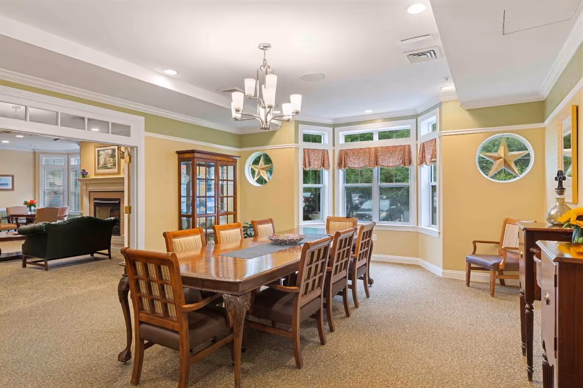 A bright dining room with a large wooden dining table surrounded by eight chairs. The room has beige walls with white trim and a bay window with patterned valances. There is a chandelier hanging above the table, a wooden cabinet with glass doors, and decorative star wall hangings. In the background, a living area with a fireplace and seating is visible.