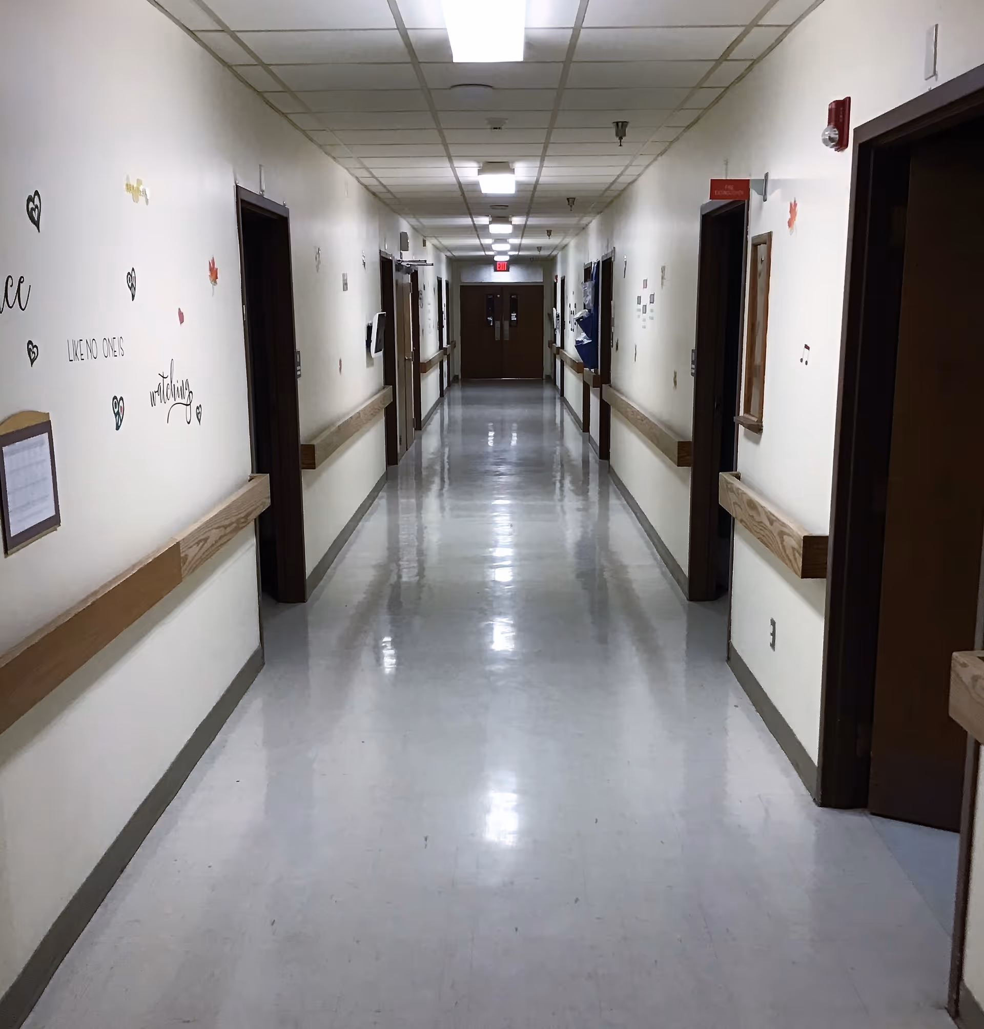 Long polished interior hallway in a senior living facility with handrails, decorated walls, and doors on both sides leading to an exit.