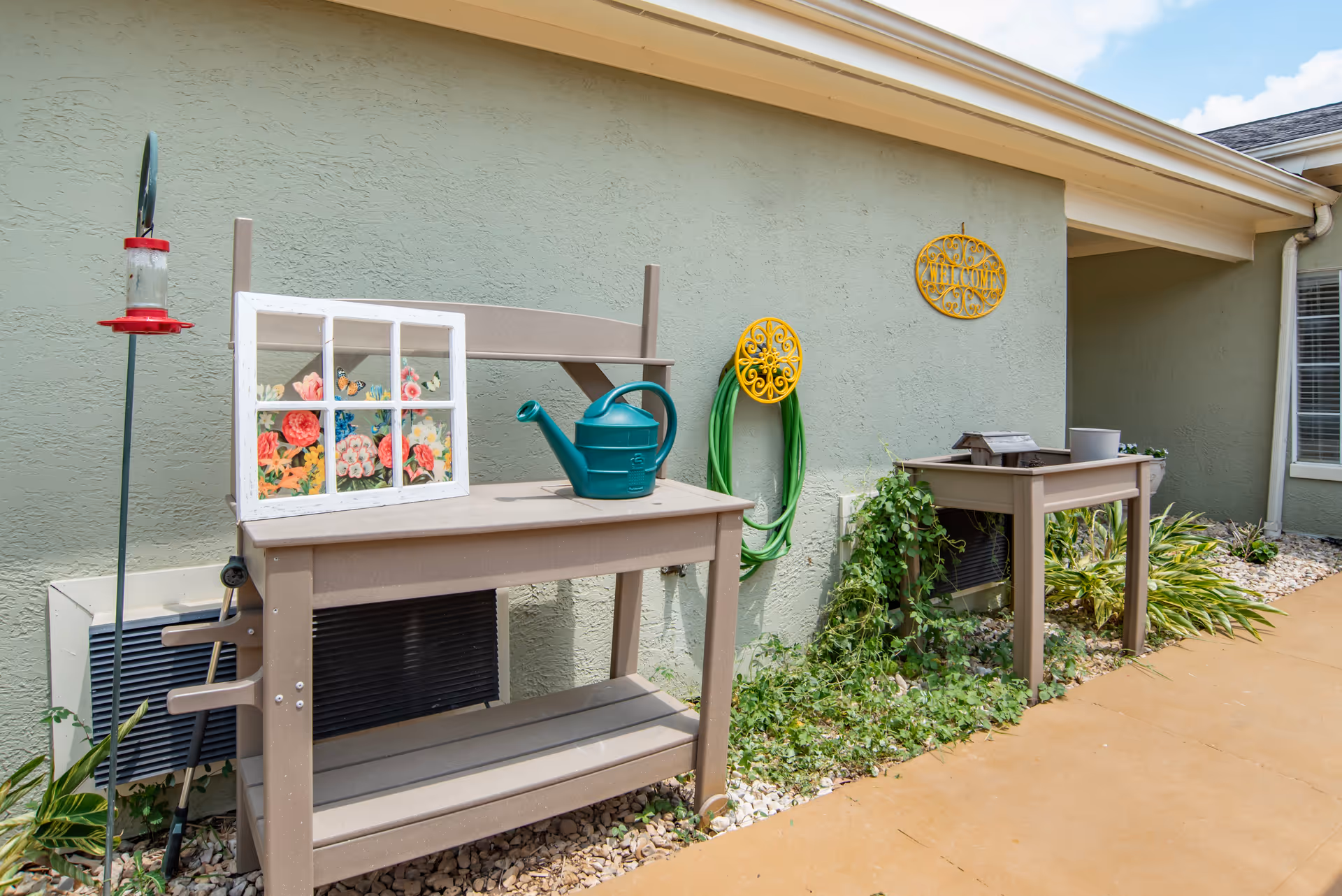 Outdoor gardening area with two beige potting benches against a light green wall. One bench has a green watering can and a decorative window frame with painted flowers. A green garden hose is coiled on the wall with a yellow holder. There are plants and rocks along the base of the wall, and a hummingbird feeder is visible on the left side.