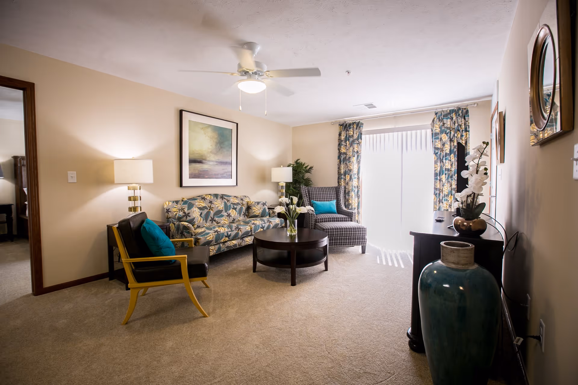 A cozy living room in a retirement facility featuring a floral patterned sofa, a checkered armchair with a turquoise pillow, a black chair with a turquoise pillow, a round wooden coffee table with a vase of flowers, two table lamps, a ceiling fan, floral curtains, a large window with vertical blinds, and decorative items including a large blue vase and a wall painting.
