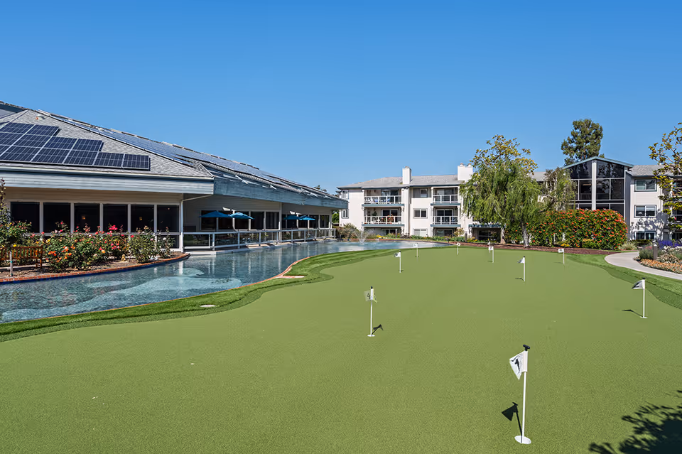 Outdoor putting green with multiple small flags in front of a senior living facility building under a clear blue sky. The building has solar panels on the roof, large windows, and a water feature with flowers and umbrellas along the side.
