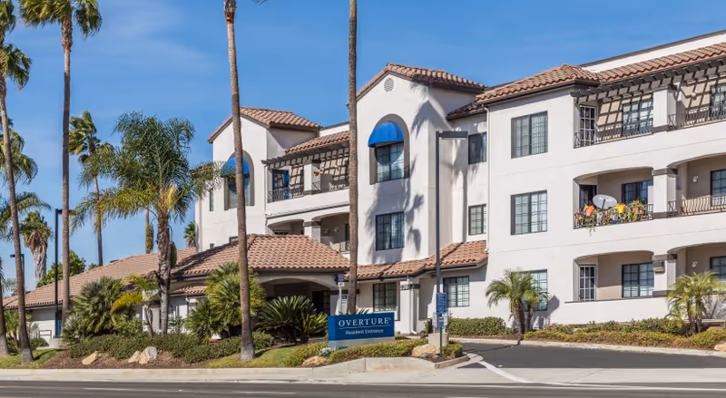 Front exterior of the Overture San Marcos senior living building with palm trees, balconies, and a sign at the driveway.