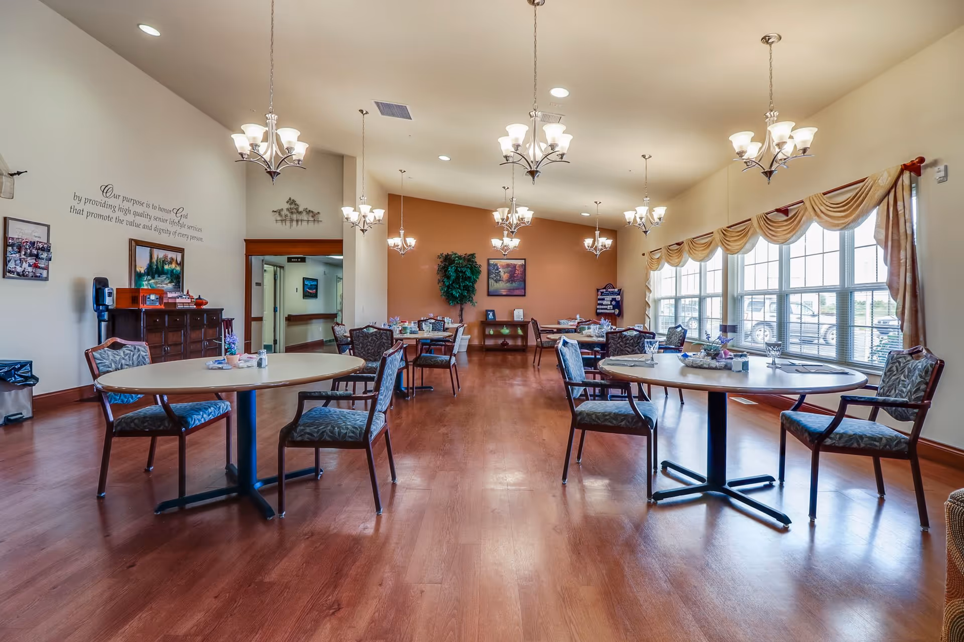 A spacious dining room with multiple round tables and chairs arranged neatly. The room has wooden flooring, large windows with draped curtains allowing natural light to fill the space, and several chandeliers hanging from the ceiling. There is a decorative plant and framed artwork on the far wall, along with a sideboard and a water dispenser on the left side.