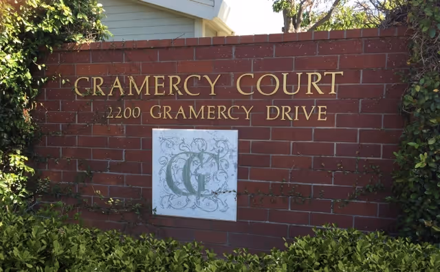 A brick sign with gold lettering that reads 'Gramercy Court 2200 Gramercy Drive' with a decorative emblem below the text, surrounded by green bushes and some ivy on the sides.
