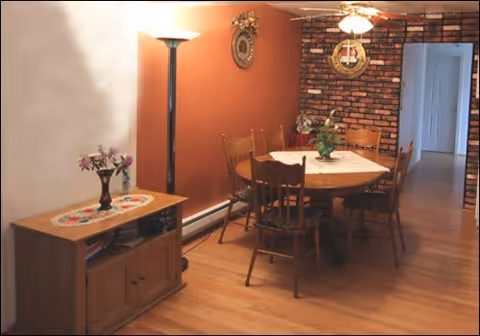 A dining area with a round wooden table covered with a lace tablecloth and surrounded by six wooden chairs. A green plant centerpiece is on the table. The room has hardwood floors and a brick accent wall with a clock and decorative items. A floor lamp and a wooden cabinet with a vase of flowers are also visible.