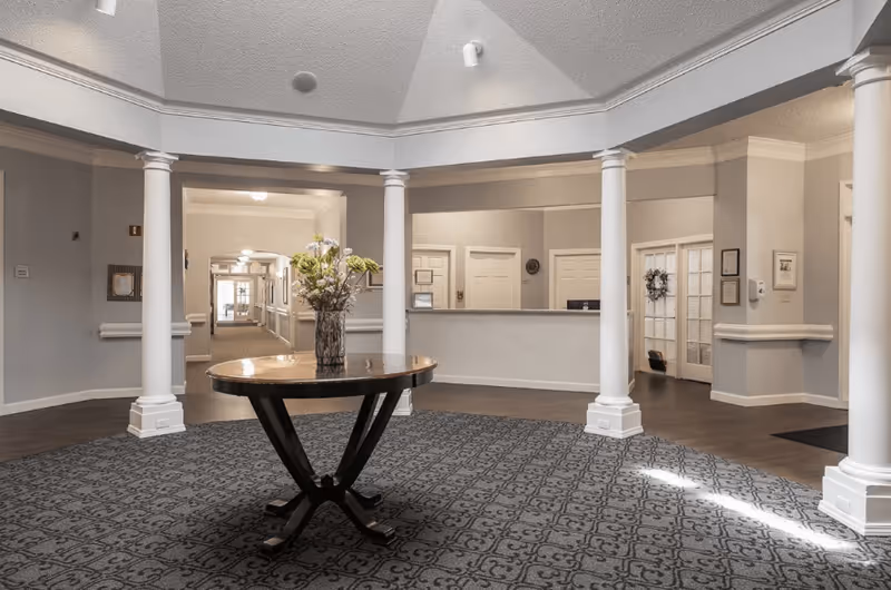 Interior view of a senior living facility lobby area with a round wooden table holding a vase of flowers in the center, surrounded by white columns and a patterned carpet. In the background, there is a reception desk and hallway with doors and framed pictures on the walls.