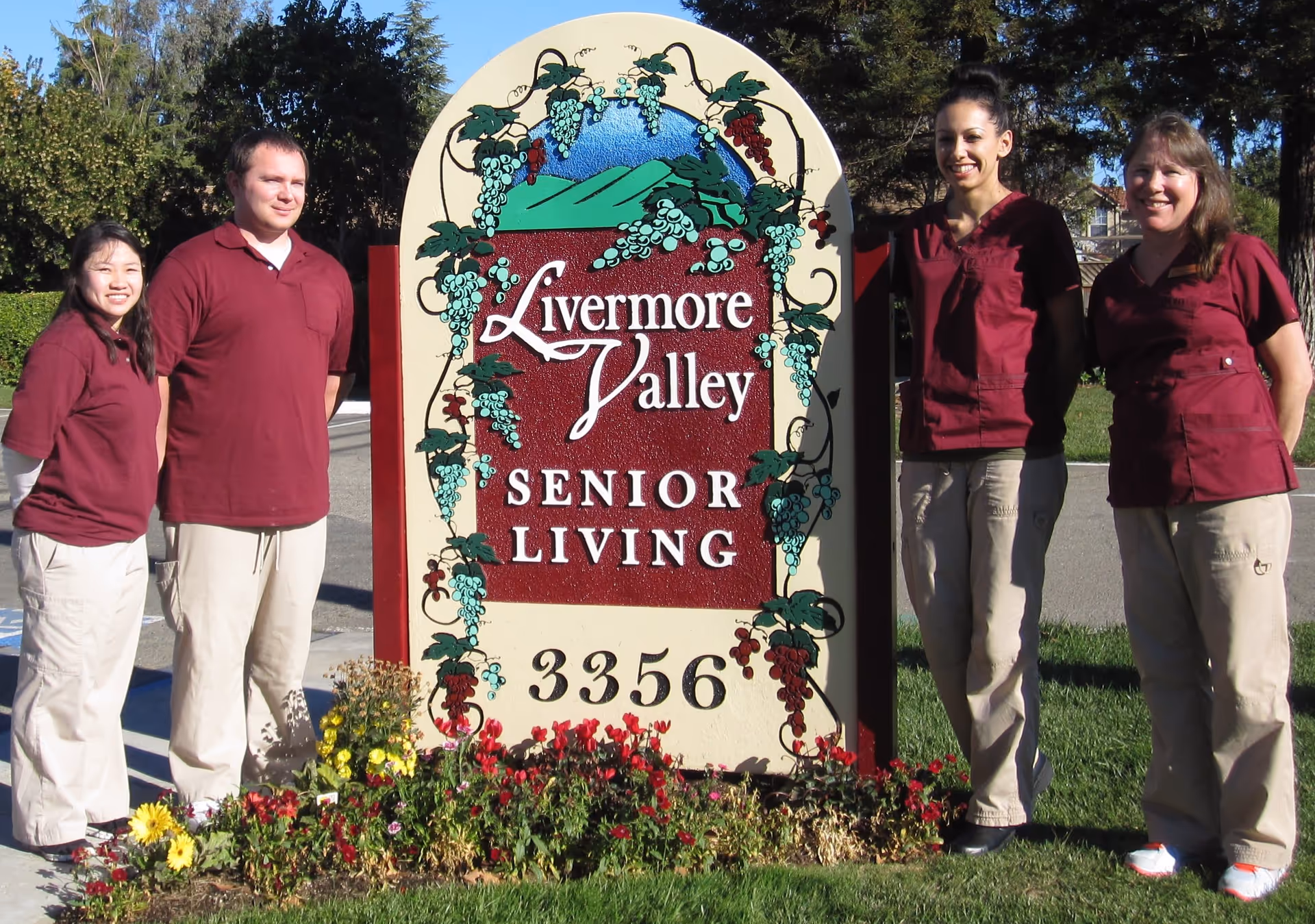 Four staff members in uniforms stand beside a decorative sign that reads "Livermore Valley Senior Living" with flowers planted at its base.