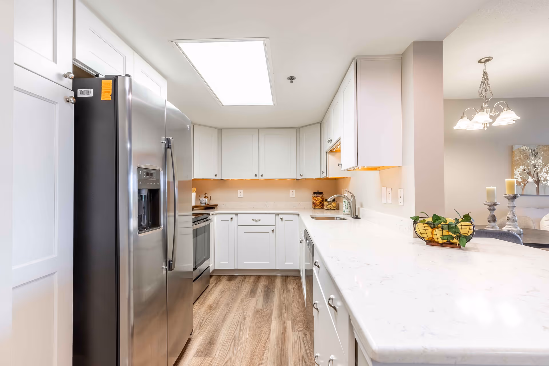 Bright and modern kitchen with white cabinets, stainless steel refrigerator and oven, marble countertops, wooden flooring, and a basket of fruit on the counter. The kitchen opens into a dining area with a chandelier and decorative candles.