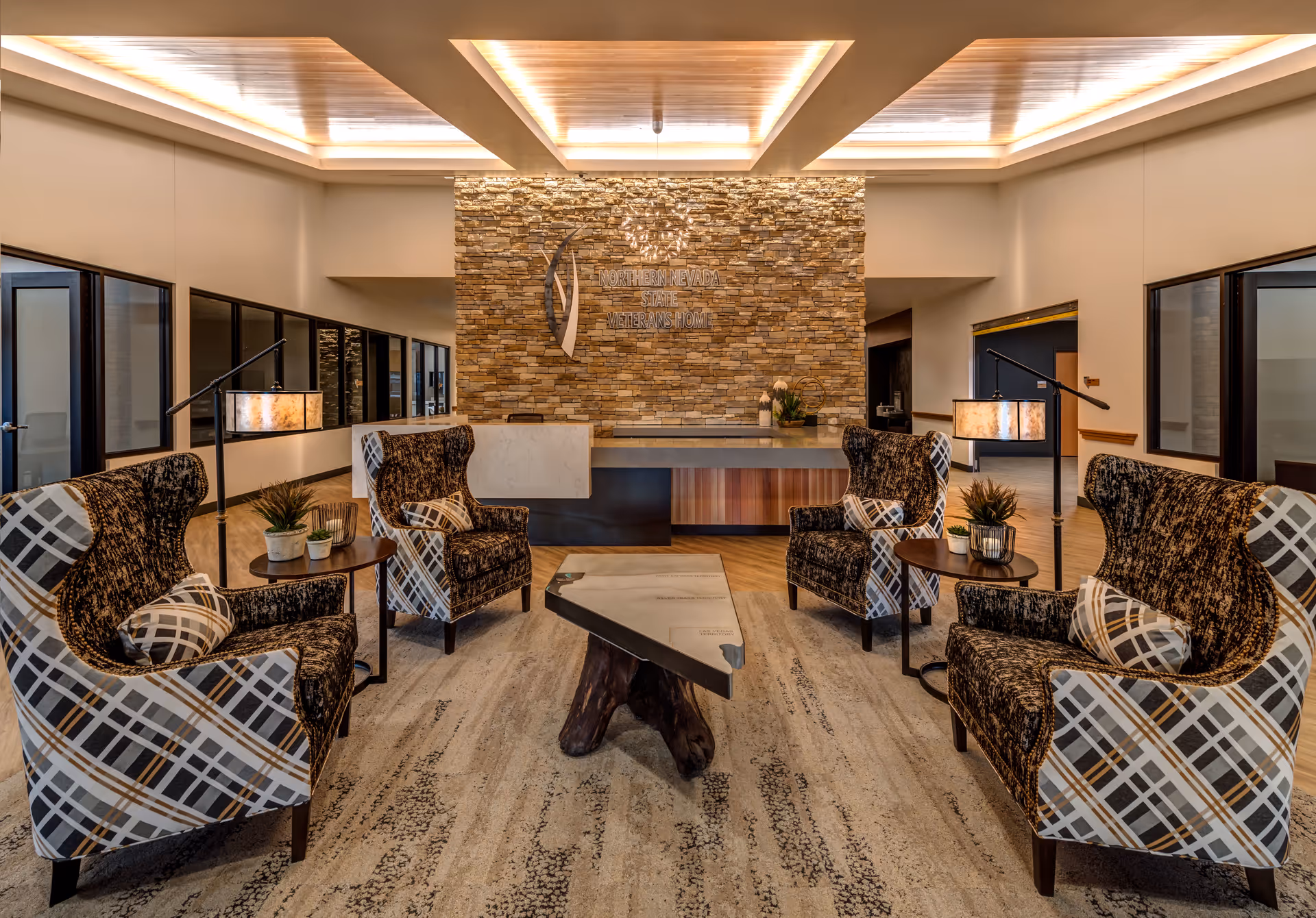 A modern and welcoming lobby area with four patterned armchairs arranged around a unique wooden coffee table. The background features a stone accent wall with the sign 'Northern Nevada State Veterans Home' and a reception desk. The space is well-lit with recessed ceiling lights and has large windows along the sides.