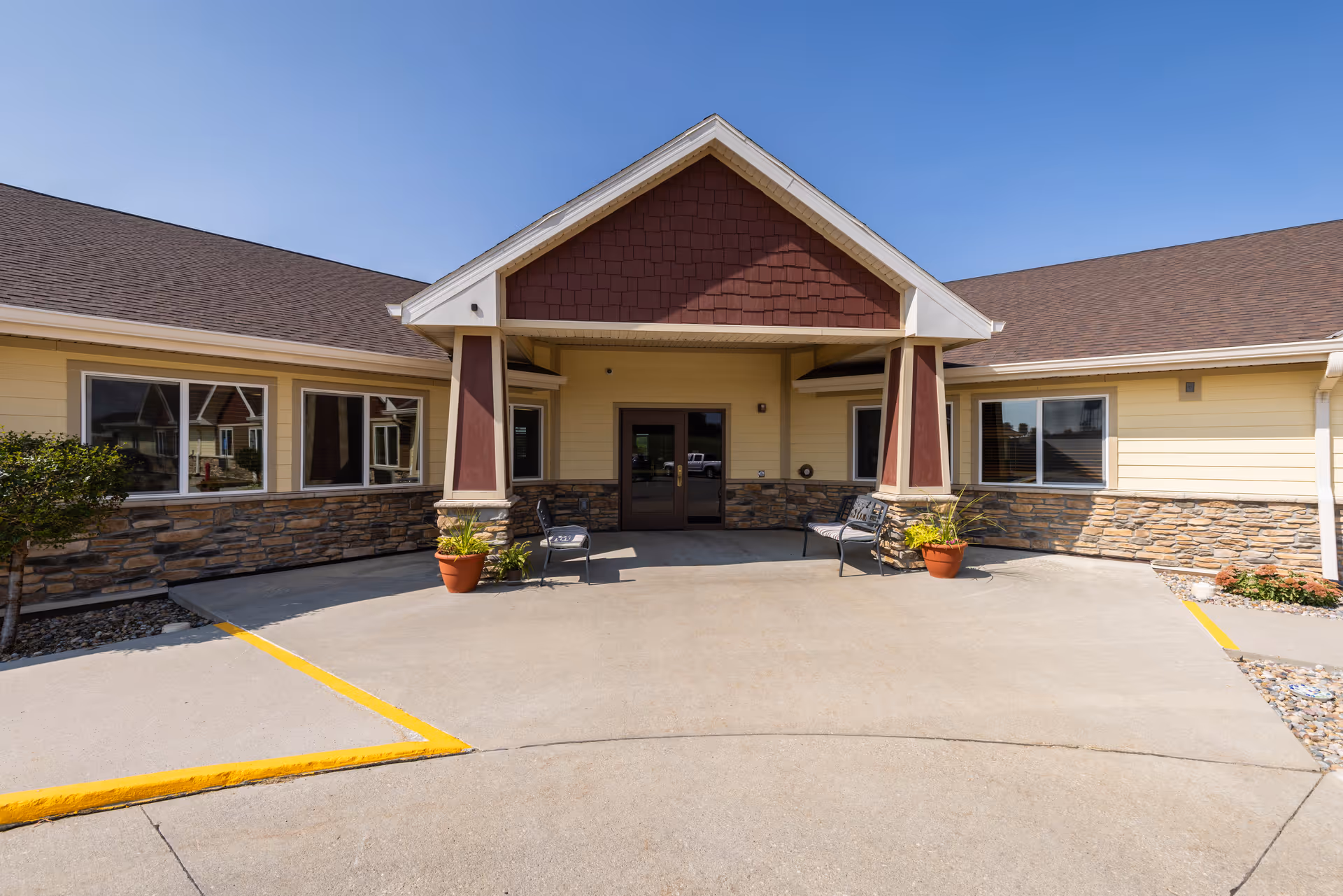 Front entrance of a senior living facility with a covered porch supported by two columns. The building exterior features stone and yellow siding with large windows. Two benches and potted plants are placed near the entrance under a clear blue sky.