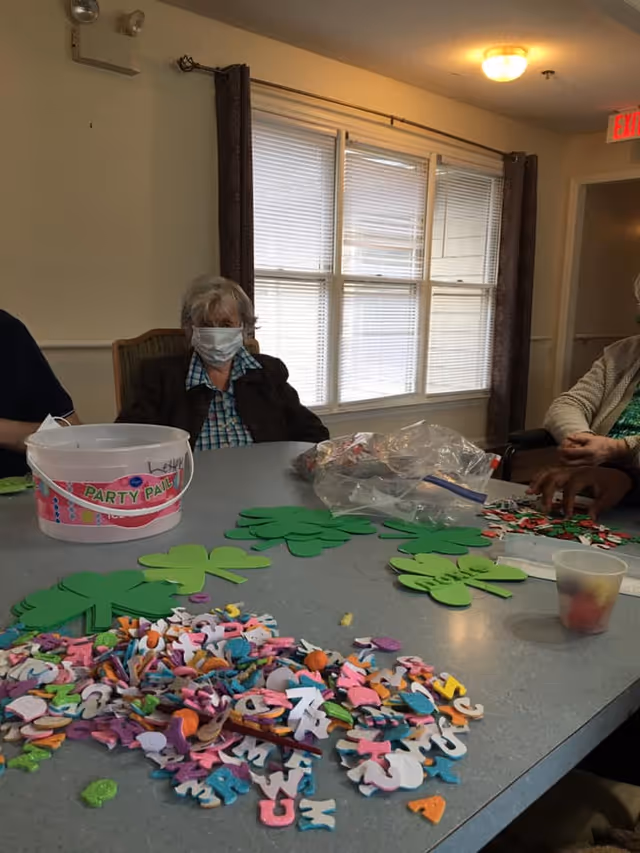 A group of elderly individuals sitting around a table in a well-lit room with a window in the background. The table is covered with colorful craft materials including foam letters, green shamrock shapes, and a plastic bucket labeled 'Party Pail'. One elderly woman is wearing a face mask and a dark jacket.