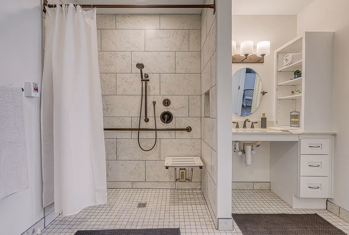 Accessible bathroom with a walk-in shower featuring a handheld showerhead, grab bars, and a fold-down shower seat. Adjacent to the shower is a vanity with a sink, an oval mirror, open shelving with towels and decorative items, and three drawers. The bathroom has light-colored tiles and walls, with dark floor mats.
