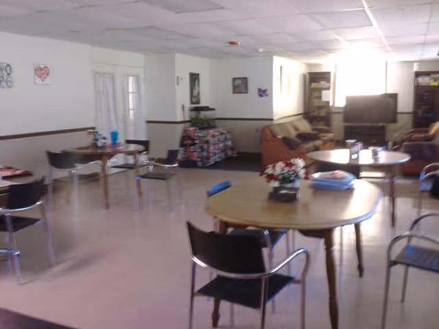 A common area in a senior living facility with several round wooden tables and black chairs arranged around the room. There are couches and a television in the background, along with a fish tank on a table covered with a colorful cloth. The walls are decorated with framed pictures and artwork.