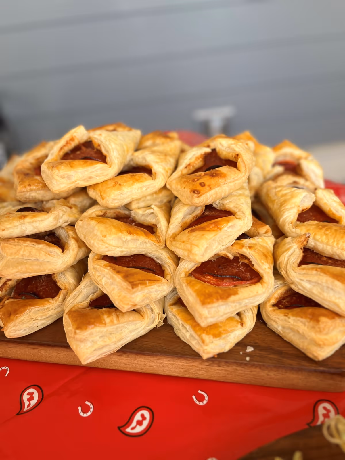 A pile of golden puff pastry appetizers with pepperoni centers on a wooden serving board atop a red patterned tablecloth.