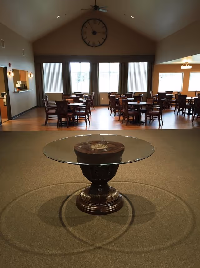 Interior view of a dining area in a senior living facility with multiple round wooden tables and chairs arranged on a wooden floor. In the foreground, there is a round glass table with a decorative wooden base placed on a carpeted floor. Large windows with vertical blinds and curtains allow natural light into the room. A large wall clock is mounted on the wall above the windows.
