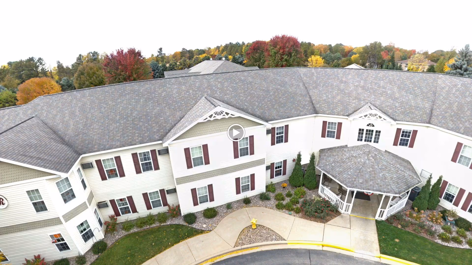 Aerial view of Renaissance by Rennes - Marinette senior living facility showing a two-story building with white siding and maroon shutters, a covered entrance with a gazebo-style roof, landscaped garden beds, and a curved driveway with a yellow fire hydrant. Trees with autumn foliage are visible in the background.