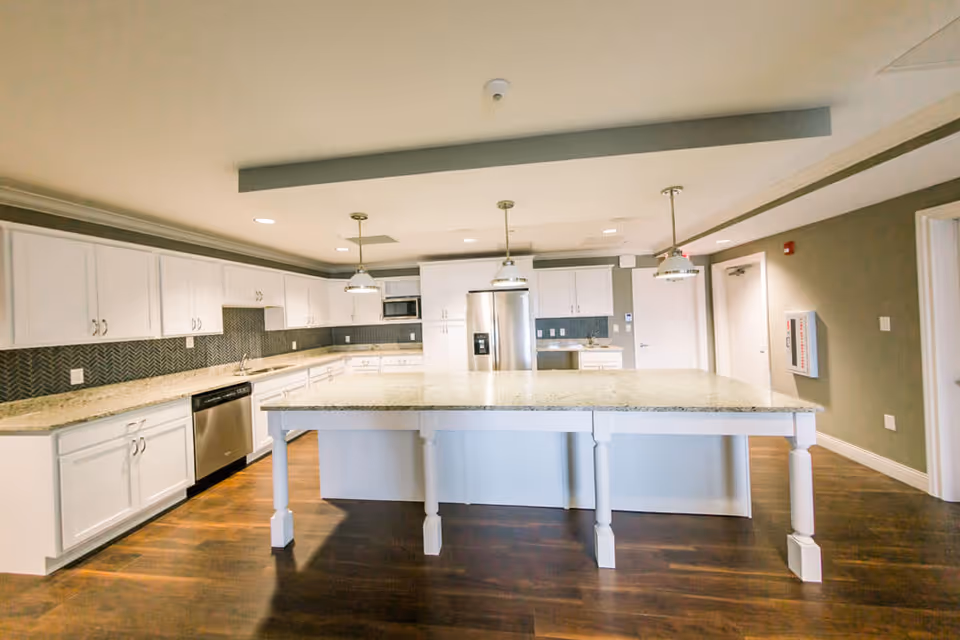 A spacious modern kitchen with white cabinetry, a large central island with a granite countertop, stainless steel appliances including a refrigerator and dishwasher, pendant lighting, and dark wood flooring.