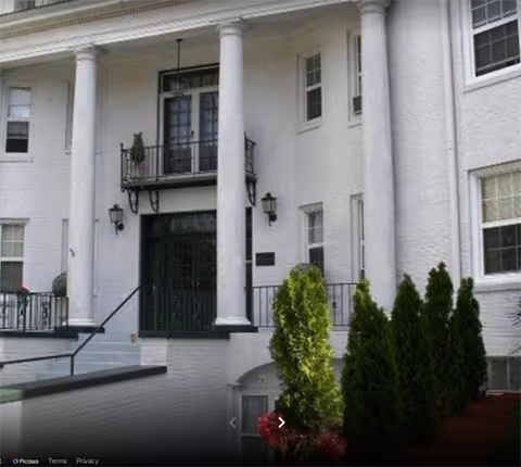 Front exterior view of a white building with large columns, black doors, and multiple windows. There are shrubs and greenery in front of the building.