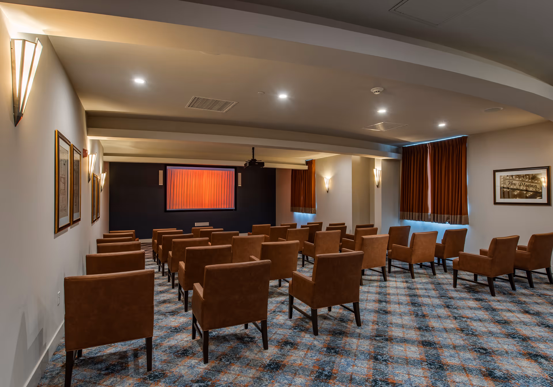 A small theater or screening room with rows of brown upholstered chairs facing a screen with a red curtain. The room has patterned carpet, wall sconces for lighting, framed pictures on the walls, and windows with brown curtains.