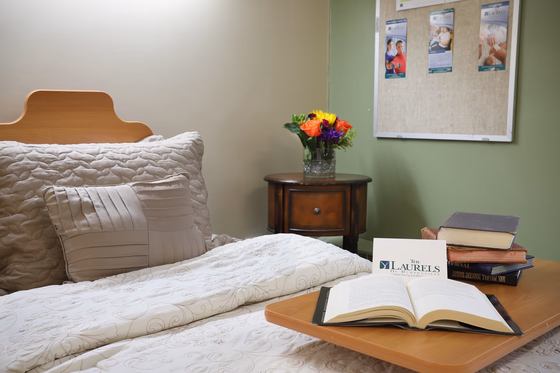A cozy bedroom setup with a neatly made bed featuring a quilted beige comforter and matching pillows. Next to the bed is a wooden nightstand with a glass vase holding a colorful bouquet of flowers. On the bed is a wooden tray holding an open book, a stack of closed books, and a card with the logo and name 'The Laurels of Hudsonville'. A bulletin board with flyers is mounted on the green wall behind the nightstand.