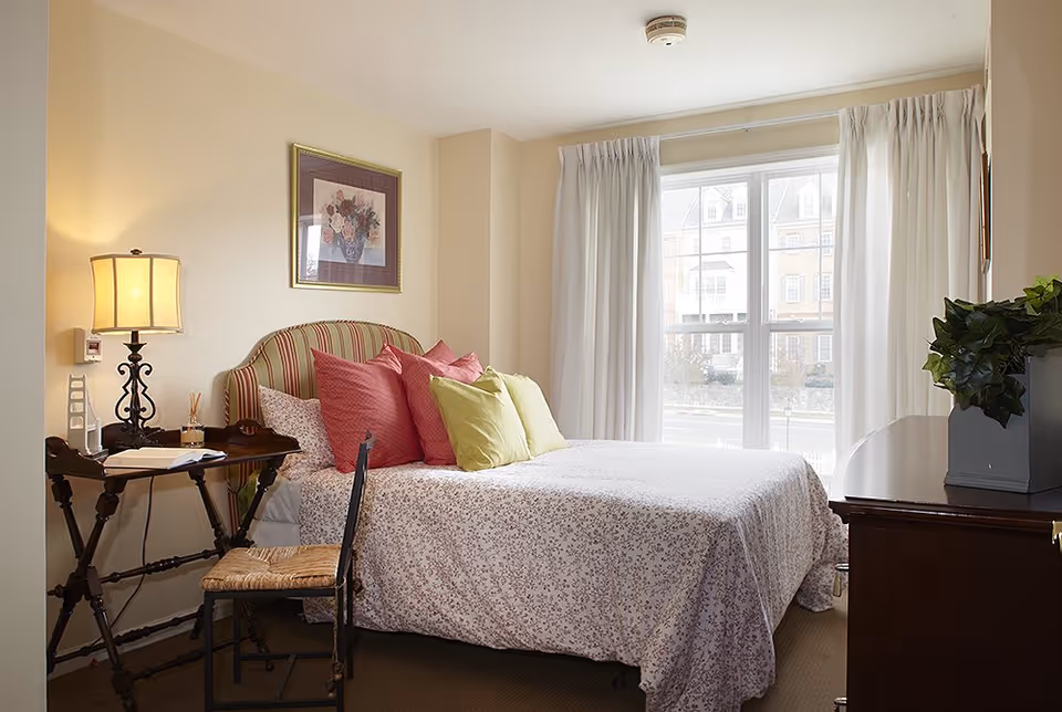A cozy bedroom with a bed covered in a floral bedspread and multiple pillows in shades of pink and green. There is a wooden side table with a lamp and some items on it, a wooden chair with a woven seat, a framed floral picture on the wall, and a window with white curtains letting in natural light. A dark wooden dresser with a potted plant is also visible.