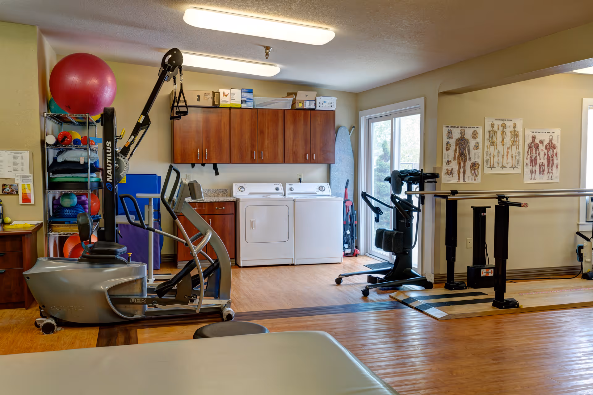Physical therapy room with exercise equipment including a Nautilus machine, parallel bars for walking support, anatomical charts on the wall, a shelf with exercise balls and other therapy tools, and a washer and dryer in the background.