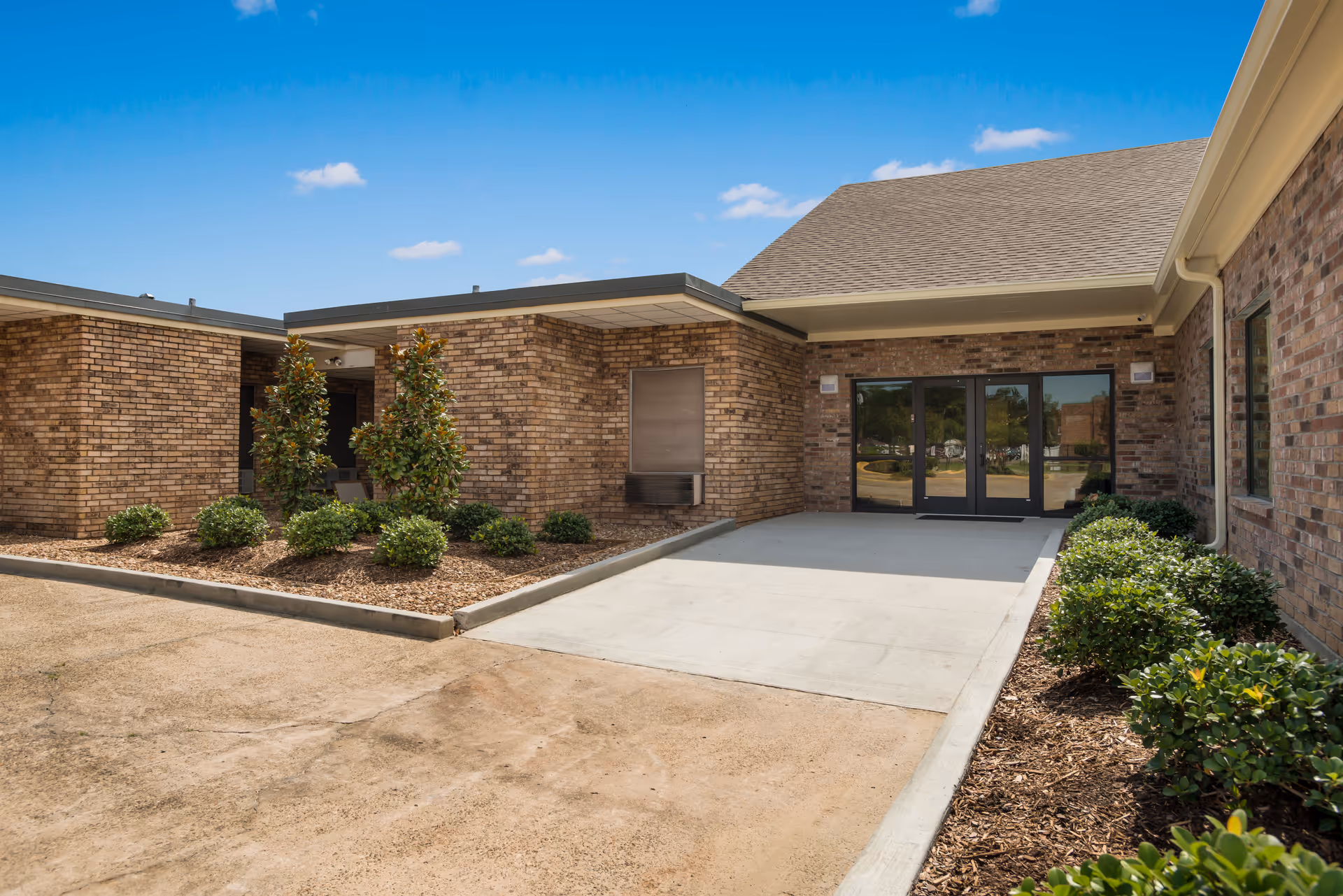 Exterior view of a brick building with a concrete pathway leading to double glass doors under a covered entrance. The area is landscaped with small bushes and trees under a clear blue sky.