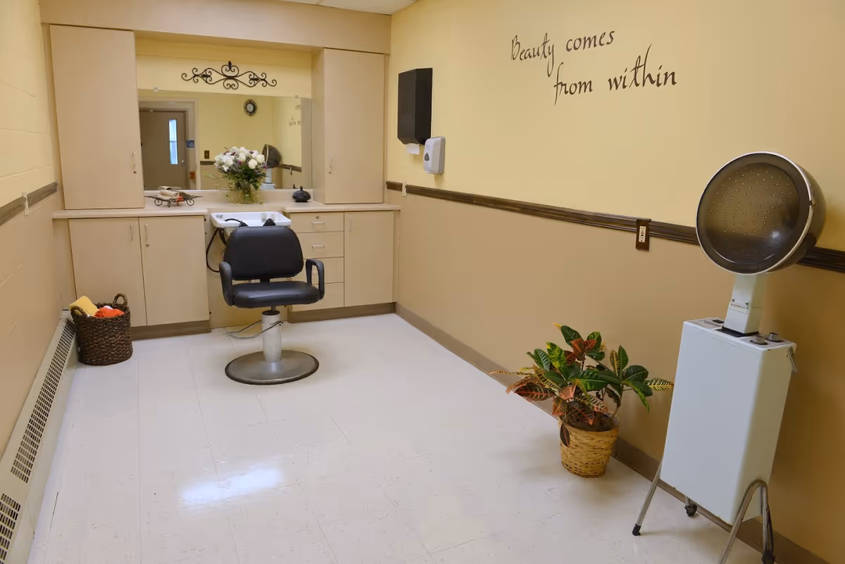 Interior salon room with a styling chair and sink, hooded hair dryer, cabinets, and a potted plant beneath a wall decal reading "Beauty comes from within."