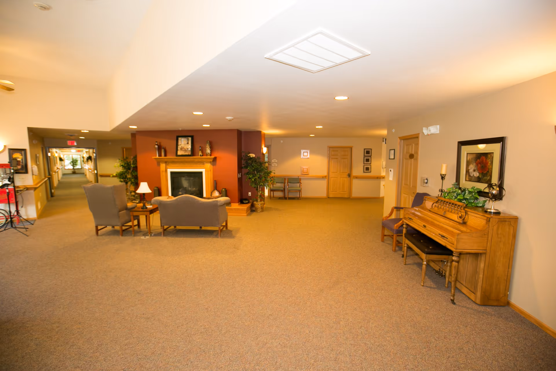 A spacious senior living facility common area with beige carpet and walls. There is a wooden piano with a bench and decorative items on top to the right. In the center, there is a seating area with two armchairs and a loveseat around a wooden coffee table, positioned in front of a fireplace with a wooden mantel and red accent wall. The hallway extends to the left with framed pictures on the walls and plants placed near the seating area and along the hallway.