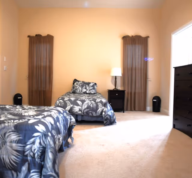 Bright bedroom with two twin beds in matching floral bedding, a nightstand with a lamp, a dresser, and two narrow curtained windows.