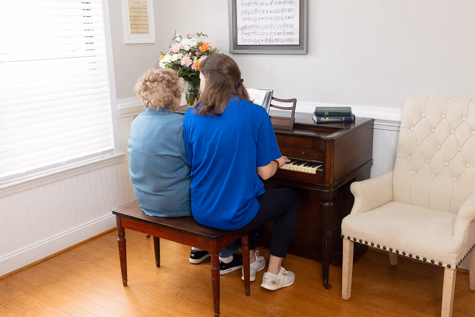 An elderly woman and a younger woman sitting side by side on a wooden bench playing a piano in a well-lit room with a large window, a vase of flowers on the piano, framed sheet music on the wall, and a beige upholstered armchair nearby.