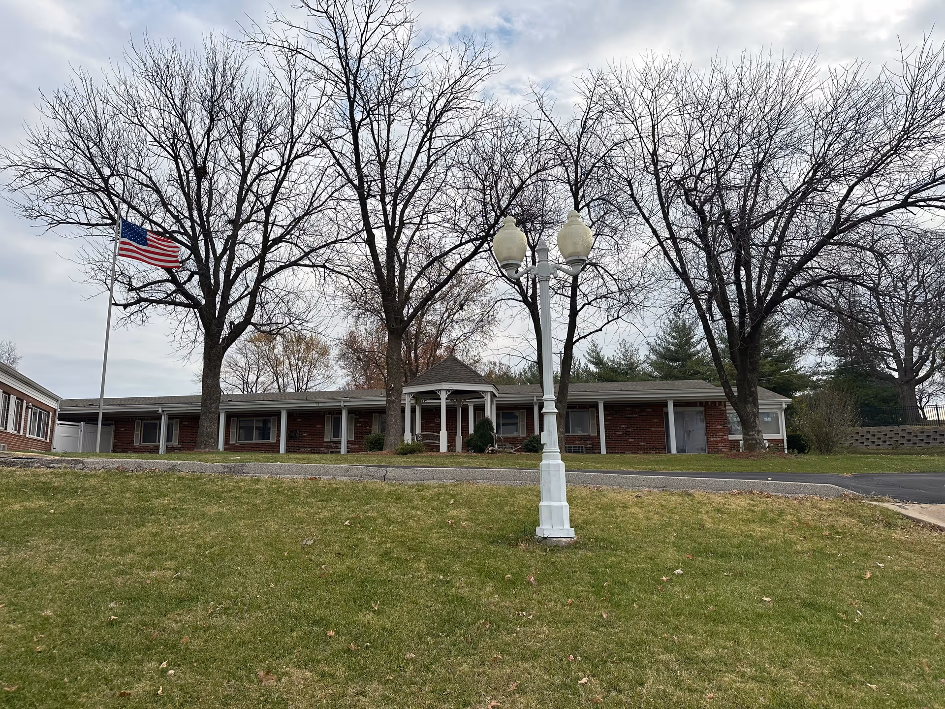 Exterior view of a single-story brick building with a covered porch area, surrounded by leafless trees and a green lawn. There is an American flag on a flagpole to the left and a white street lamp in the foreground. The sky is cloudy.