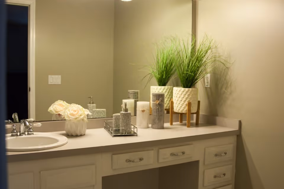 A bathroom countertop with a sink, decorated with two white artificial flowers in a vase, two potted green plants in white textured pots on a wooden stand, two candles, and a soap dispenser. A large mirror is mounted on the wall behind the countertop, reflecting the items and part of the room.