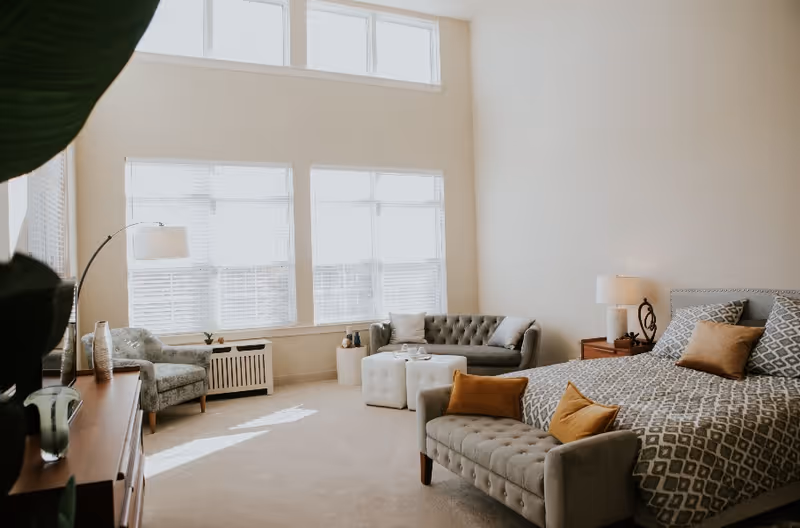 A bright and spacious bedroom with large windows letting in natural light. The room features a bed with patterned bedding and multiple pillows, a cushioned bench at the foot of the bed, a gray tufted sofa with pillows, a small armchair, a wooden dresser with decorative items, a floor lamp, and a bedside table with a lamp.