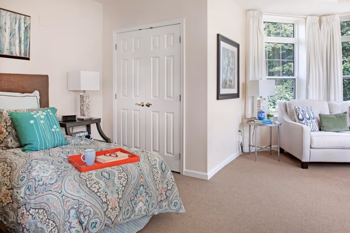 Sunlit bedroom with a patterned bed and tray, bedside tables, and a seating area with a sofa by large bay windows.