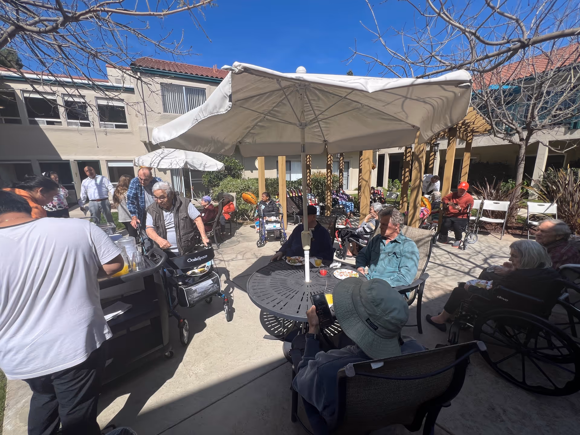 A group of elderly people sitting and socializing outdoors under large white umbrellas in a courtyard area of a senior living facility. Some individuals are in wheelchairs and others are seated on chairs around tables. Caregivers and staff are also present, assisting and interacting with the residents. The building with windows and a tiled roof surrounds the courtyard, and leafless trees are visible under a clear blue sky.