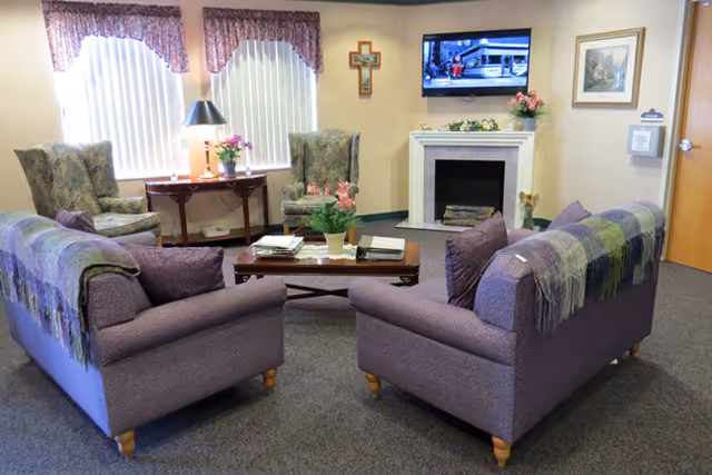 Comfortable common lounge with two purple sofas facing a coffee table, armchairs, a fireplace, and a wall-mounted TV.