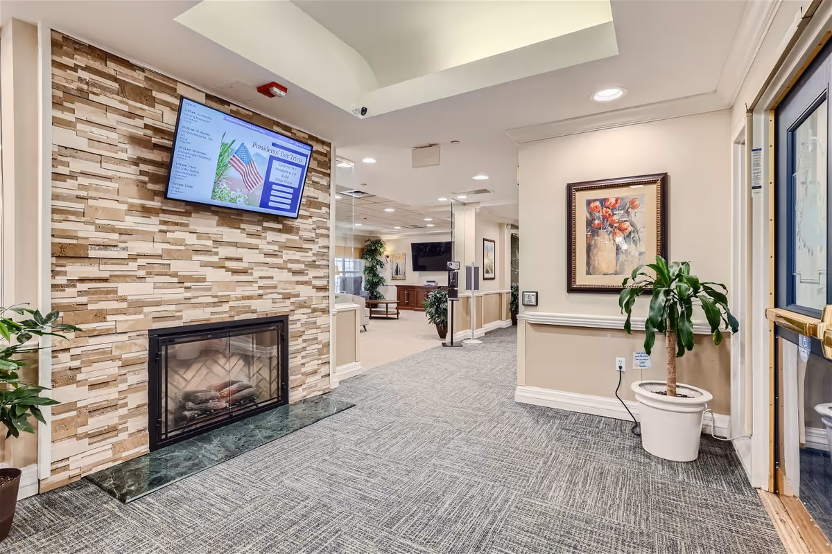 Interior view of a senior living facility hallway with a stone accent wall featuring a fireplace and a mounted flat-screen TV displaying a President's Day menu. The hallway has carpeted flooring, potted plants, framed artwork on the walls, and leads into a common area with seating and additional plants.