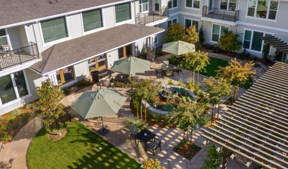 A courtyard area at Oakmont of Novato featuring outdoor seating with tables and umbrellas, surrounded by greenery, small trees, and a circular water fountain in the center. The courtyard is enclosed by a multi-story building with windows and balconies.