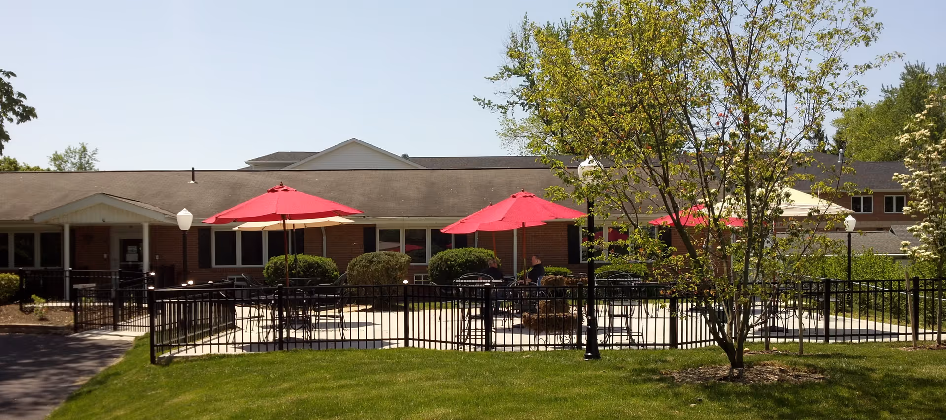 Outdoor patio area at Bethany Nursing Home with several tables shaded by red umbrellas, surrounded by a black metal fence. The building is a single-story brick structure with a sloped roof. There are green bushes and trees around the patio, and the sky is clear and blue.