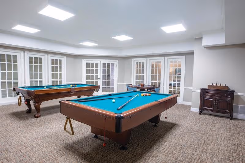 A spacious game room with two pool tables, one in the foreground and one in the background. The room has beige carpeting, white walls with a chair rail, and multiple white-framed glass doors along the walls. There is a small wooden cabinet with a chess set on top in the corner.