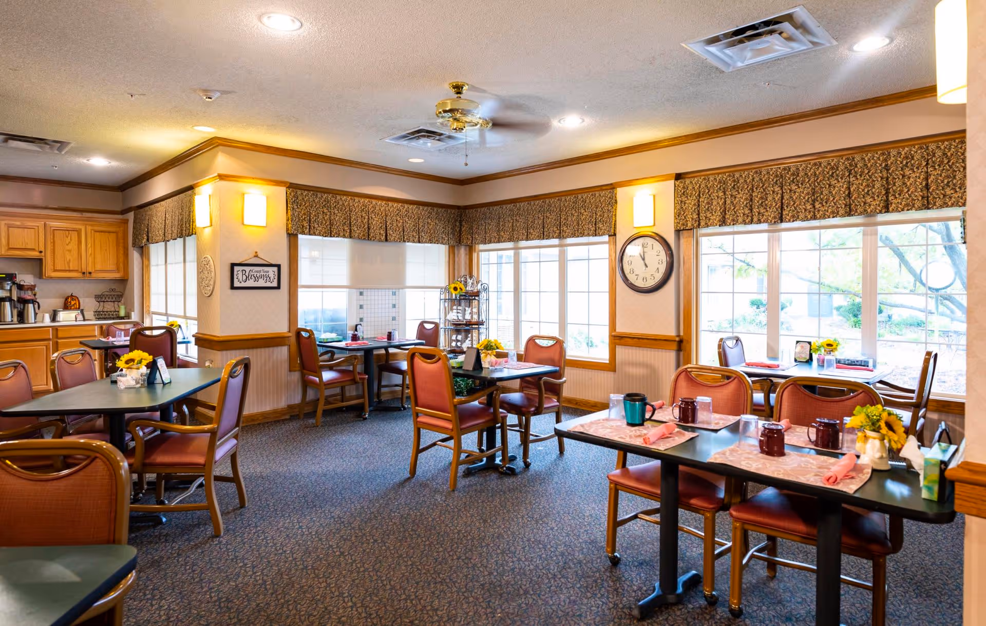 Dining room in a senior living facility with multiple set tables and chairs, large windows, and a serving counter.