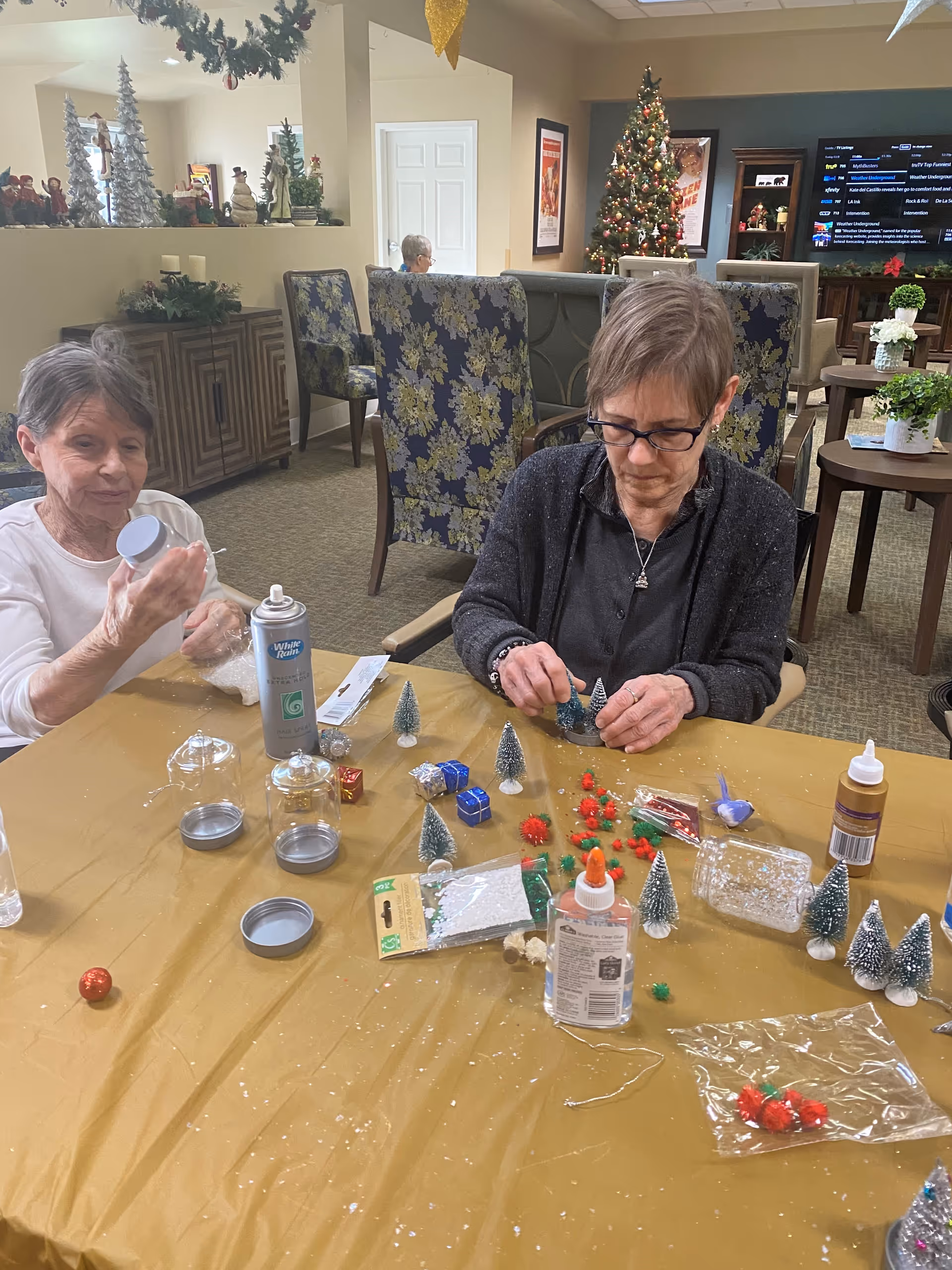 Two elderly women sitting at a table covered with a yellow tablecloth, engaging in a holiday craft activity with miniature Christmas trees, small gift boxes, glue, and decorative items. The room is decorated for Christmas with a Christmas tree, holiday figurines, and garlands. In the background, there are patterned chairs, a TV, and other residents seated.