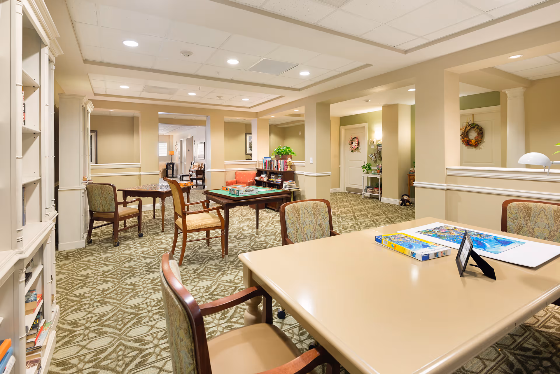 A well-lit common area in a senior living facility with multiple tables and chairs arranged for activities. One table has a puzzle and a box on it, while another table has a board game set up. The room features patterned carpet, beige walls with white trim, and decorative wreaths on the walls. Bookshelves and plants add to the cozy atmosphere.