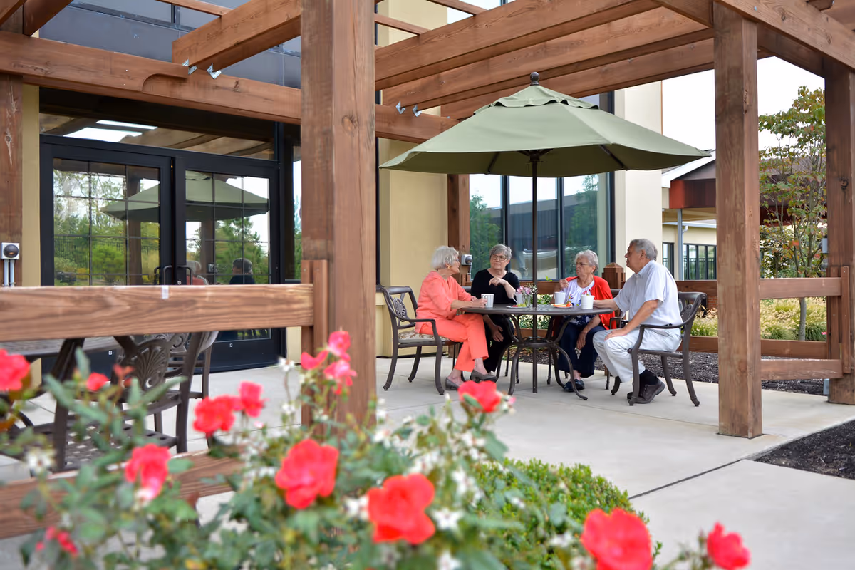 Four elderly people sitting around a round table under a green patio umbrella, engaged in conversation outside a building with large windows and wooden pergola structure. Red flowers and greenery are visible in the foreground.