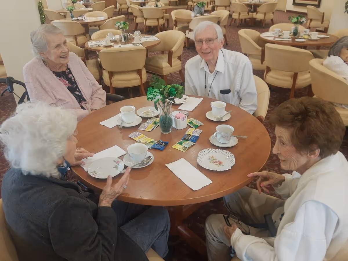 Four elderly residents seated around a round table enjoying tea in a communal dining room.
