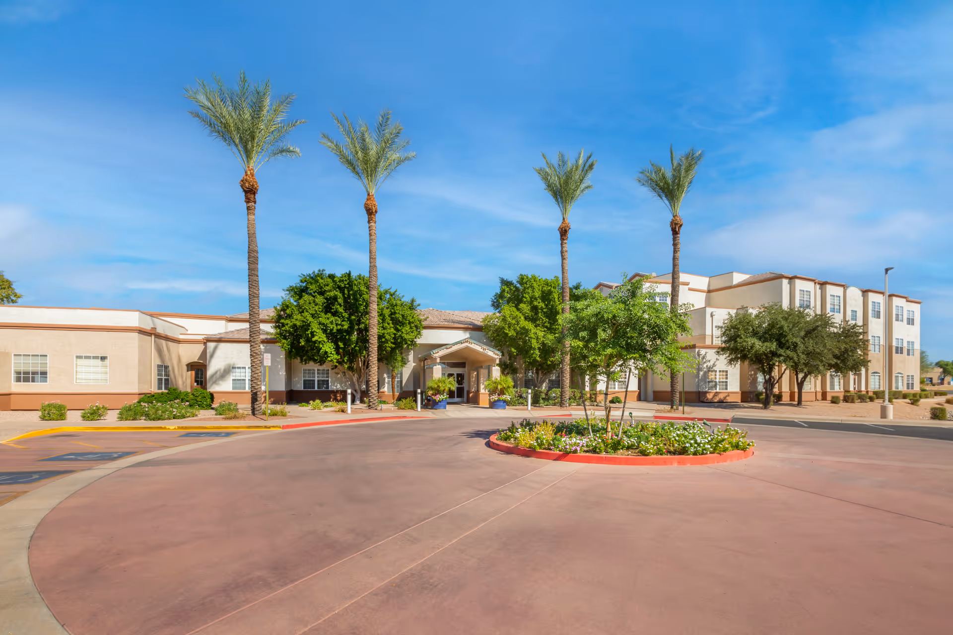 Exterior view of Brookdale Baywood senior living facility featuring a circular driveway with a landscaped center island, tall palm trees, and a beige multi-story building under a clear blue sky.