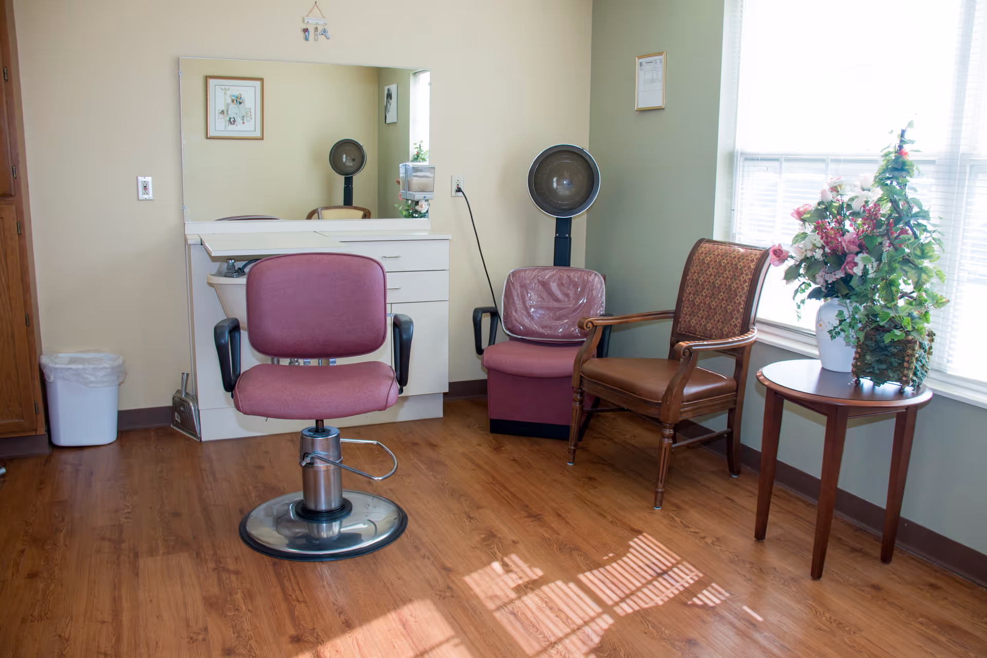A small salon-style room with a styling chair in front of a mirror, a hooded hair dryer, two waiting chairs, and a side table with a flower arrangement.